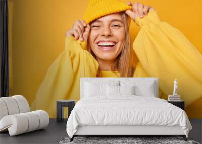 Happy playful young woman smiles broadly shows white teeth keeps hands on hat dressed in casual jumper expresses sincere emotions being in good mood isolated over yellow background. Joy concept Wall mural