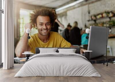 Happy positive African American college student with cheerful cute smile using wireless internet connection on laptop computer at coffee shop while looking for information online for research project Wall mural