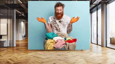 Happy red haired man with thick bristle, spreads hands, has fun after doing laundry at home, dressed in white dirty t shirt, poses over blue background. People, emotions and domestic work concept Wall mural