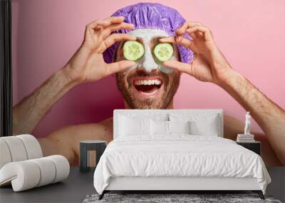 Happy relaxed young man covers eyes with two slices of cucumber, applies cosmetic mask on face, wears bathcap, stands naked against pink background. Self care, beauty and spa therapy concept Wall mural