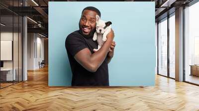 Happy satisfied smiling dark skinned man carries small pet of french bulldog breed, spend leisure time together, expresses positive emotions during photoshoot with dog, isolated over blue background Wall mural