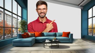 Happy smiling European male with bristle, points with index finger aside, invites you to have dinner in restaurant situated near, dressed in casual red t shirt, spectacles isolated on white background Wall mural