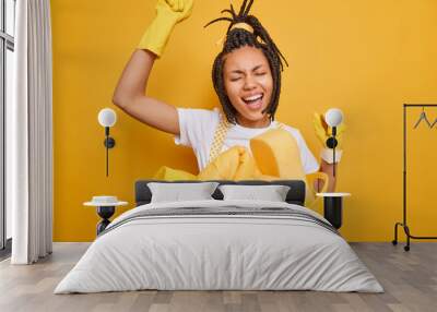 Happy woman housemaid dances carefree keeps arms raised poses near laundry basin has fun while doing household chores sings song along isolated over vivid yellow background. Housekeeping concept Wall mural