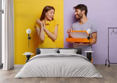 Happy young male model opens box, makes surprise for girlfriend on birthday, shows something in package, isolated over purple background. Glad lovely woman with headphones on neck, receives gift Wall mural