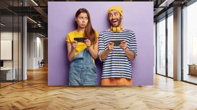 Happy young man and sad female bloggers use smartphone devices for online communication, play games, being addicted to modern technologies, stand shoulder to shoulder, express different emotions Wall mural