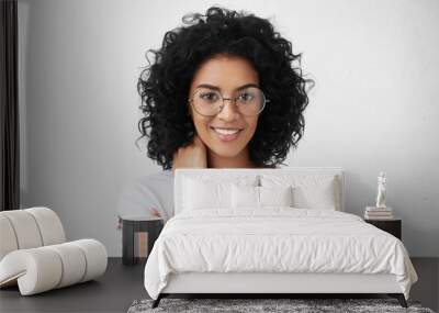 Headshot of attractive young mixed race woman wearing big spectacles and white top holding hand behind her neck and looking at camera with subtle smile during awkward conversation with some guy Wall mural