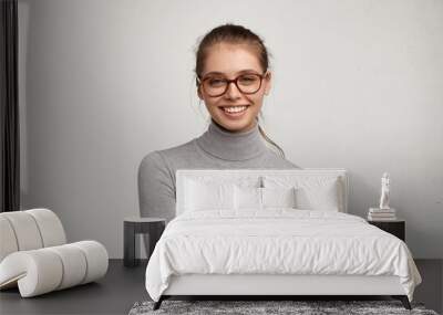 Headshot of cute student girl wearing grey turtleneck sweater and stylish eyeglasses smiling broadly, having carefree and relaxed look while resting at home after morning lectures at college Wall mural