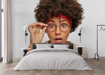 Headshot of emotive shocked young woman has bushy Afro hairstyle, has eyes popped out, gazes through spectacles, dressed in casual clothes, models indoor. Stupefaction and amazement concept. Wall mural