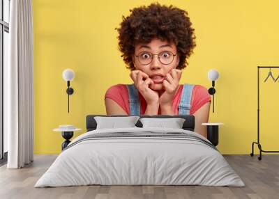 Headshot of frustrated mixed race young woman has crisp hair, bites finger nails with nervous expression, stares with eyes full of fear, wears casual clothes, stands against yellow studio wall Wall mural