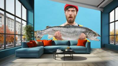 Headshot of funny emotional young bearded Caucasian fisherman in hat and raincoat holding large salmon in both hands, staring at camera with shocked look, can't believe that he caught such a big fish Wall mural