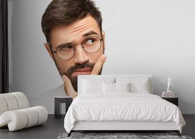 Headshot of good looking bearded young male holds chin and looks pensively aside, raises eyebrows and ponders about something, tries to find correct solution in mind, poses over white blank wall Wall mural