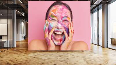 Headshot of overjoyed happy ethnic woman keeps hands on cheeks giggles positively keeps mouth opened has creative makeup smeared face with watercolor paints stands naked against rosy background Wall mural