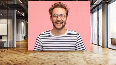 Headshot of pleased hipster has satisfied expression, curly hair and bristle, wears round transparent glasses and striped t shirt, feels glad after promotion at work, isolated over pink background Wall mural