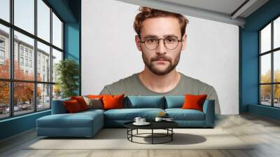 Headshot of serious bearded man with mustache and beard, wears round spectacles, has stylish hairdo, looks confidently, models in white studio, waits for partner. Young student going for classes Wall mural