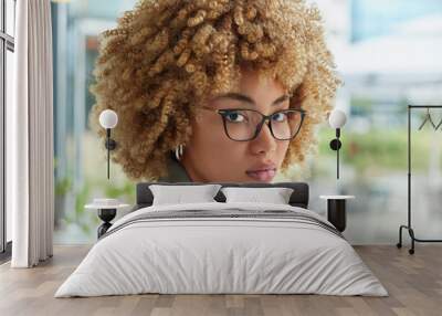 Headshot of serious successful woman with curly blonde hair keeps hand under chin wears optical spectacles and formal clothes looks attentively at camera poses outdoors against blurred background Wall mural