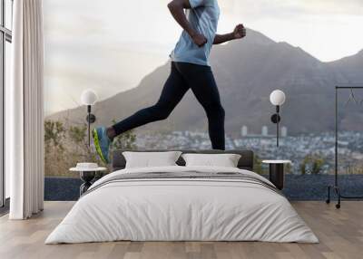 Healthy dark skinned man in action, runs along road near mountains, wears comfortable sneakers, casual clothes, has sporty body. Fast male athlete poses against sky background. Racing competition Wall mural