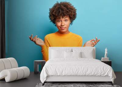 Hesitant dark skinned woman has puzzled expression shrugs shoulders looks confused dressed in casual clothes isolated over blue background. Indecisive Afro American female makes doubt gesture Wall mural