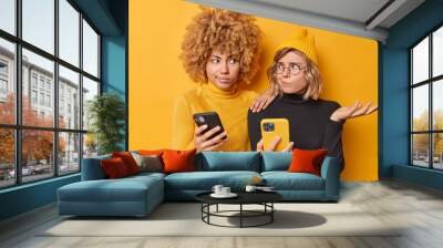 Hesitant young two women look uncertain at each other use modern mobile phones use new smartphone application wear black and yellow turtleneck hat pose indoor dont know how to use new gadget Wall mural