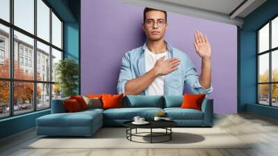 Honest serious hipster guy swears to do something, keeps hand on chest, gestures with palm, promises something, wears spectacles and denim shirt, swears indoor, isolated over purple background Wall mural