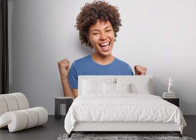 Hooray I did it. Happy young woman with curly hair clenches fists rejoices success achieves prize dressed in casual blue t shirt isolated over white background. Lucky female model becomes champion Wall mural