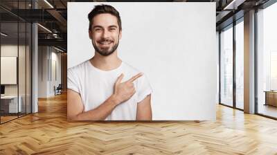 Horizontal portrait of bearded cheerful man has smile, wears casual white t shirt, indicates with fore finger at copy space for your promotional text or advertisment, isolated over concrete wall Wall mural