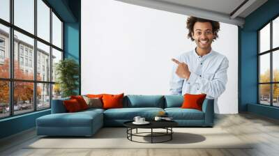 Horizontal portrait of dark-skinned handsome man having broad smile wearing formal white shirt posing against white background pointing with index finger at white copy space for your advertisment Wall mural