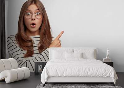 Horizontal shot of beautiful young Caucasian female with surprised expression, points at upper right corner, dressed in striped sweater, wears round spectacles, isolated over white background Wall mural