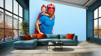 Horizontal shot of caring father gives piggyback to small child, play interesting superhero game, wear special costumes, pose against blue background with free space, have great impact on world Wall mural