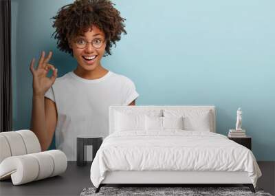 Horizontal shot of cheerful Afro American woman makes okay gesture, agrees with good proposal, wears spectacles and white casual t shirt, says incredible offer, isolated over blue background Wall mural