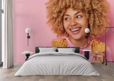 Horizontal shot of cheerful blonde woman smiles broadly shows white teeth holds two heart shaped waffles concentrated aside happily enjoys eating sweet tasty food poses against pink wall copy space Wall mural