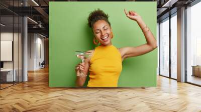 Horizontal shot of cheerful curly haired woman dances carefree wears yellow t shirt and earrings has pink manicure holds glass of cocktail isolated over vivid green background has fun on party. Wall mural