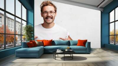 Horizontal shot of cheerful male with happy look points at white caual t shirt with copy space for your print or design. Bearded male nerd advertises new outfit, poses against blank white background Wall mural