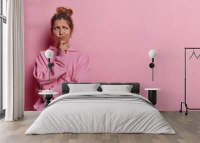 Horizontal shot of contemplative thoughtful European woman keeps hand on chin has expression of intrigue and uncertainty purses lips dressed in casual hoodie isolated over pink background copy space Wall mural