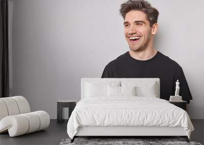 Horizontal shot of handsome cheerful man looks happily away smiles broadly wears casual black t shirt isolated over white background with blank copy space being in good mood laughs at something Wall mural