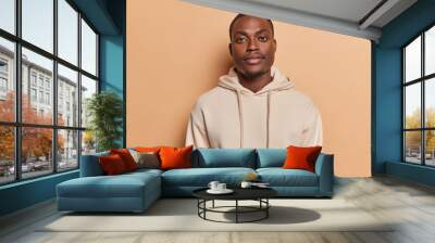 Horizontal shot of handsome dark skinned African man with small beard full lips big eyes looks directly at camera dressed in casual sweatshirt poses against brown background. Human face expressions Wall mural