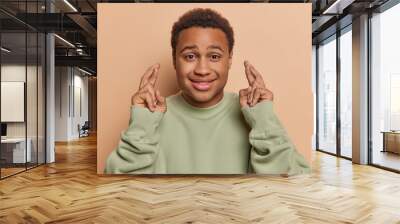 Horizontal shot of handsome dark skinned man wishing something holds fingers crossed and praying anticipating good news dressed in casual pullover isolated over brown background. Body language concept Wall mural