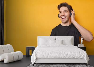 Horizontal shot of happy adult man dressed in casual black t shirt listens music via headphones enjoys favorite audio track isolated over yellow background blank copy space for your promotion Wall mural