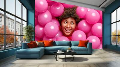 Horizontal shot of happy curly haired adult woman smiles joyfully and looks away celebrates graduation party poses among inflated air balloons cheers over holiday has fun. Celebration concept Wall mural