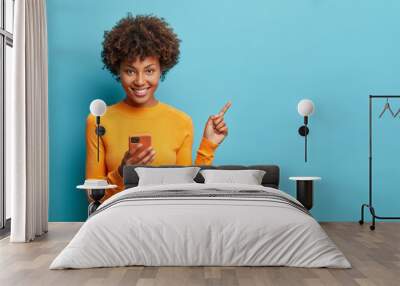 Horizontal shot of happy dark skinned Afro American woman enjoys mobile communication and modern technologies poses against blue background points away on free space for your advertising content Wall mural