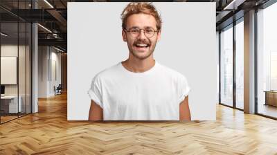 Horizontal shot of happy hipster male with toothy smile, wears casual white t shirt and glasses, being in good mood after unforgettable journey with girlfriend, isolated on white background. Wall mural