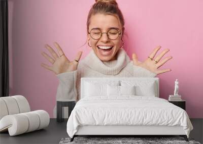 horizontal shot of happy woman with combed hair raises palms wears round spectacles and white sweate Wall mural