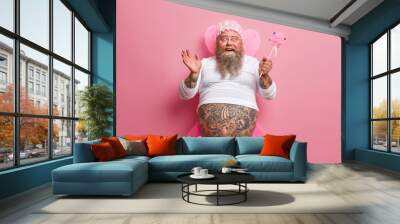 Horizontal shot of joyous funny man with thick beard and big fat belly, plays fairy on costume party, chills during spare time, sends positive vibes, works as animator, isolated over rosy background Wall mural