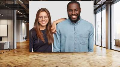 Horizontal shot of mixed race couple stand closely to each other, being in good mood, collaborate for common work, have good relationships, isolated over white background. Interracial partners Wall mural