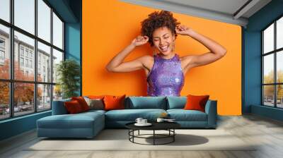 Horizontal shot of optimistic happy Afro American woman raises arms smiles broadly has upbeat mood enjoys disco party poses against vivid orange background. Women fashion style emotions concept Wall mural