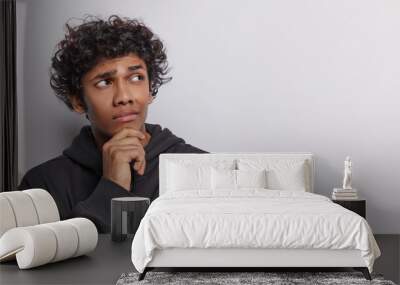 Horizontal shot of pensive curly haired teenage man keeps hand on chin concentrated aside thinking about interesting offer wears casual black sweatshirt isolated over white background copy space Wall mural