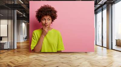 Horizontal shot of pleasant looking Afro American woman holds chin and looks pensively away, thinks about great plan, has new ideas in mind, wears bright green t shirt, poses over rosy wall copy space Wall mural