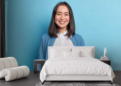 Horizontal shot of pretty Asian woman with dark hair smiles pleasantly looks directly at camera has toothy smile wears white shirt and jumper isolated over blue background. Emotions concept. Wall mural