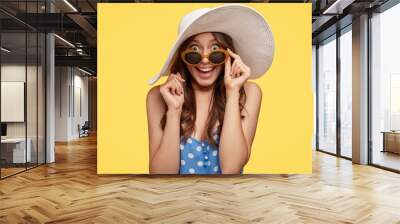 Horizontal shot of pretty attractive woman with dark hair, looks with surprise and happiness, keeps hand on rim of shades, models in summer outfit over yellow background. Beauty and emotions concept Wall mural