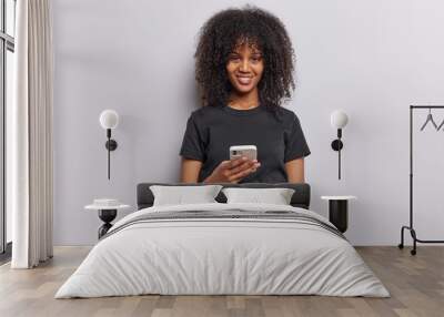 horizontal shot of pretty curly haired woman smiles positively receives messages on smartphone dress Wall mural