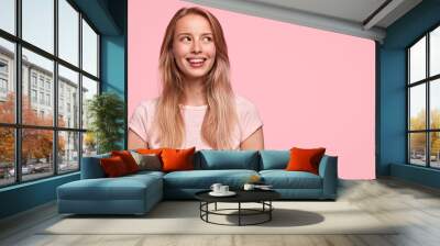 Horizontal shot of pretty happy female with broad smile, being in high spirit, dreams or imagines something pleasant, stands against pink wall with copy space. People, positive emotions concept Wall mural